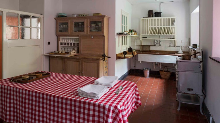 The Kitchen at Coleton Fishacre with a table in the foreground covered in a red checked tablecloth, and a stove, sink and cupboard in the background.